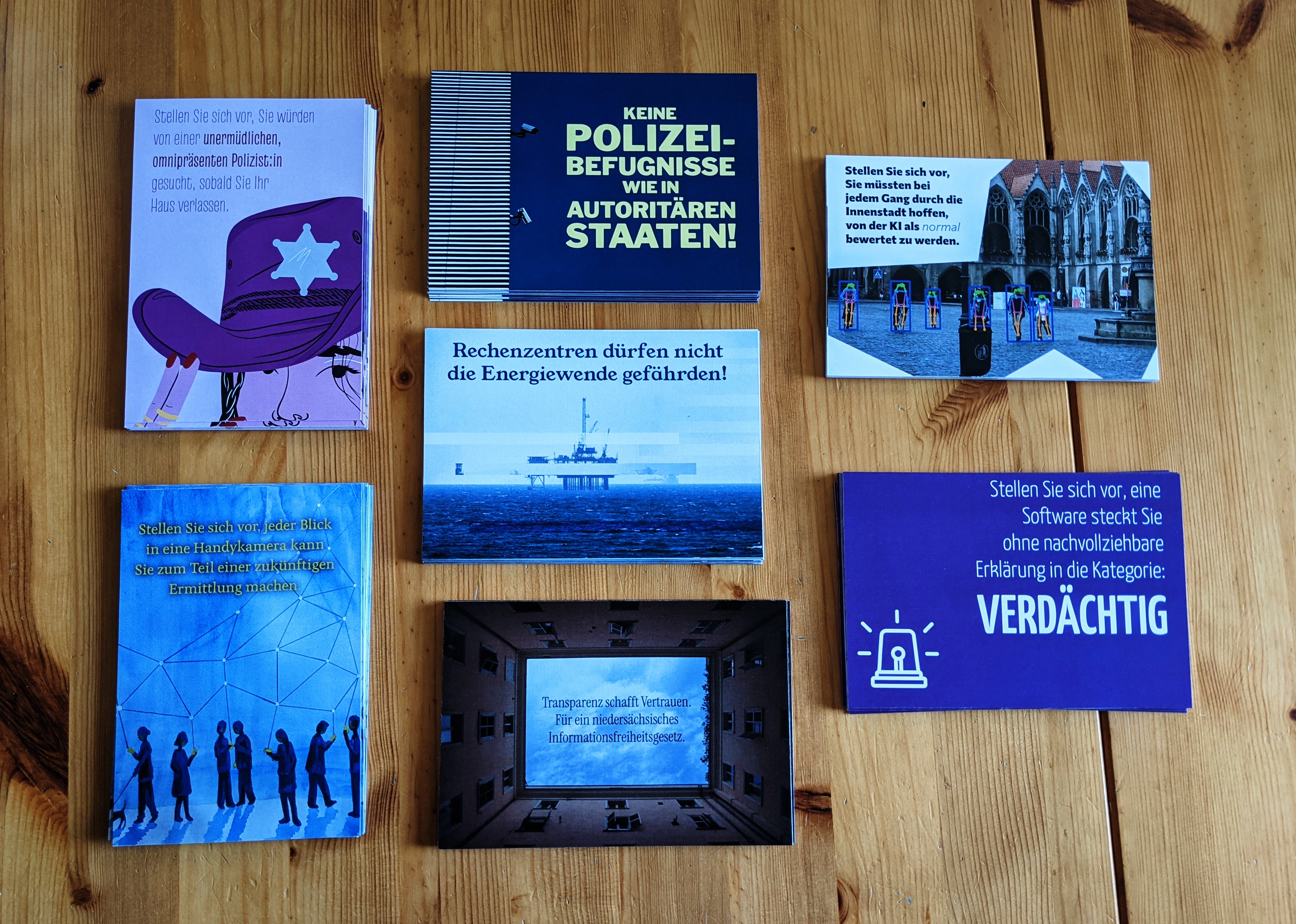 Foto von sieben Stapeln von bunten Postkarten zu Polizeigesetzen und Überwachung, Rechenzentren und Energiewende sowie Niedersächsisches Informationsfreiheitsgesetz. Auf den Postkarten sind jeweils die folgenden Sprüche und Motive: „Stellen Sie sich vor, Sie würden von einer unermüdlichen, omnipräsenten Polizist:in gesucht, sobald Sie Ihr Haus verlassen.“ (Person mit pinkem Sheriff-Hut), „Keine Polizeibefugnisse wie in autoritären Staaten!“ (zwei Überwachungskameras an einer Wand), „Stellen Sie sich vor, Sie müssten bei jedem Gang durch die Innenstadt hoffen, von der KI als normal bewertet zu werden.“ (Innenstadtszene, bei der alle Menschen mit Punktskeletten und Rahmen markiert sind), „Rechenzentren dürfen nicht die Energiewende gefährden!“ (Bohrplattform mit Glitch-Artifakten), “„Stellen Sie sich vor, jeder Blick in eine Handykamera kann Sie zum Teil einer zukünftigen Ermittlung machen“ (Menschen blicken auf ihr Handy, im Hintergrund Netzwerkgraph), „Transparenz schafft Vertrauen. Für ein niedersächsisches Informationsfreiheitsgesetz“ (Blick in den Himmel durch rechteckige Häuserfront), „Stellen Sie sich vor, eine Software steckt sie ohne nachvollziehbare Erklärung in die Kategorie: verdächtig“ (Sirenen-Icon)
